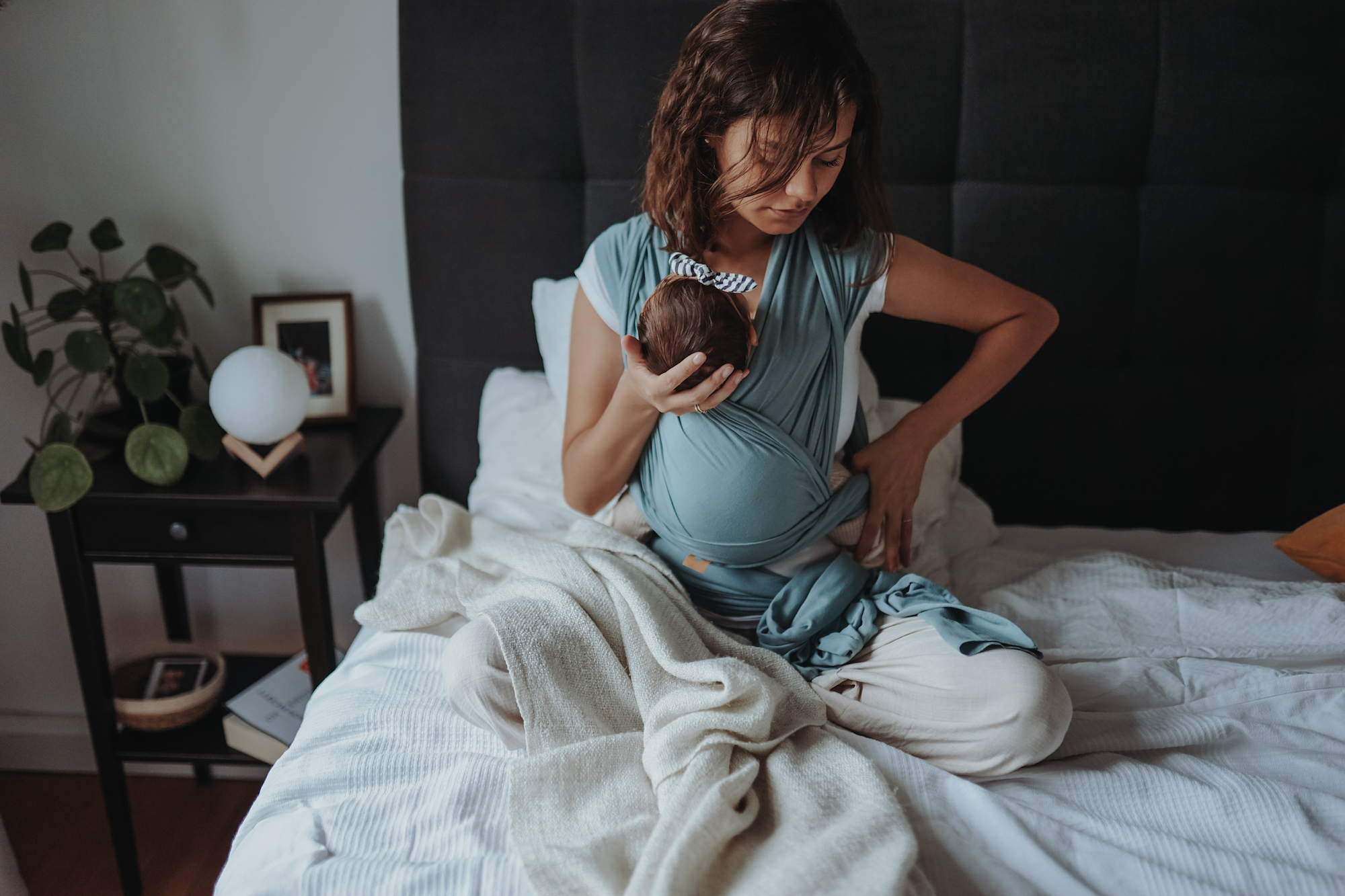 Auf diesem Bild siehst Du eine Mama, die auf dem Bett sitzt und ihr Neugeborenes im Tragetuch an sich trägt. Der Blogartikel handelt von nützlichen Tipps rund um das Thema Beckenboden.