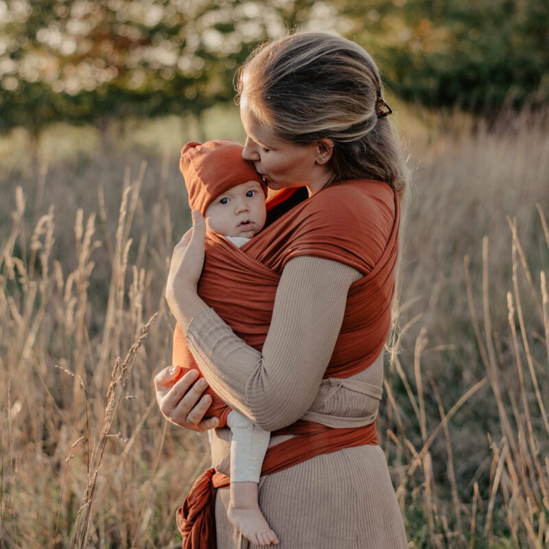 Auf diesem Bild siehst Du eine Mama mit ihrem Baby im rostfarbenen Babytragetuch